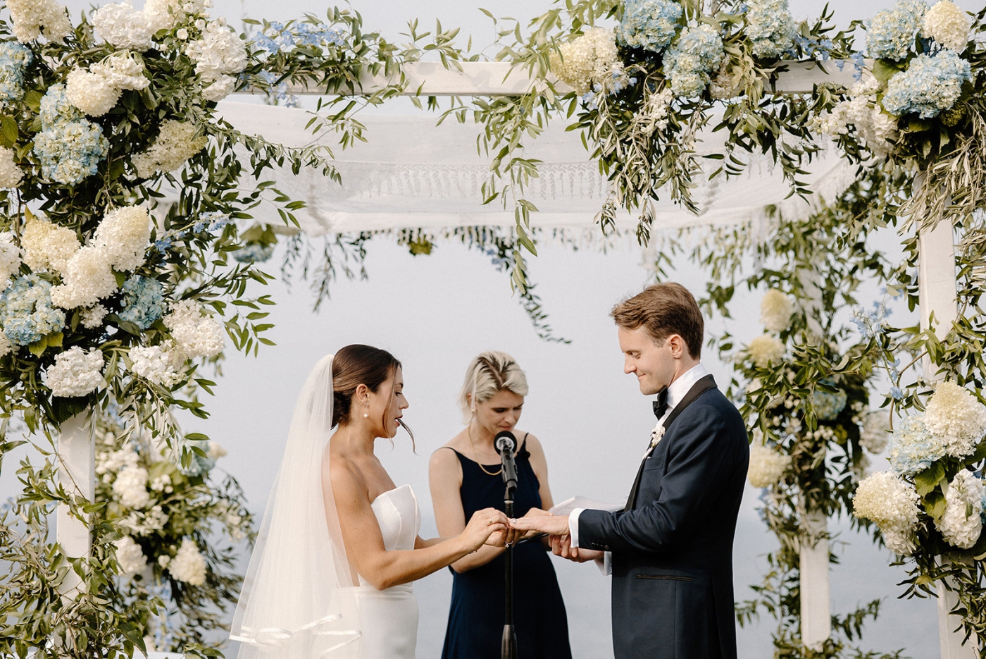 A bride and groom exchange rings beneath a large white floral installation