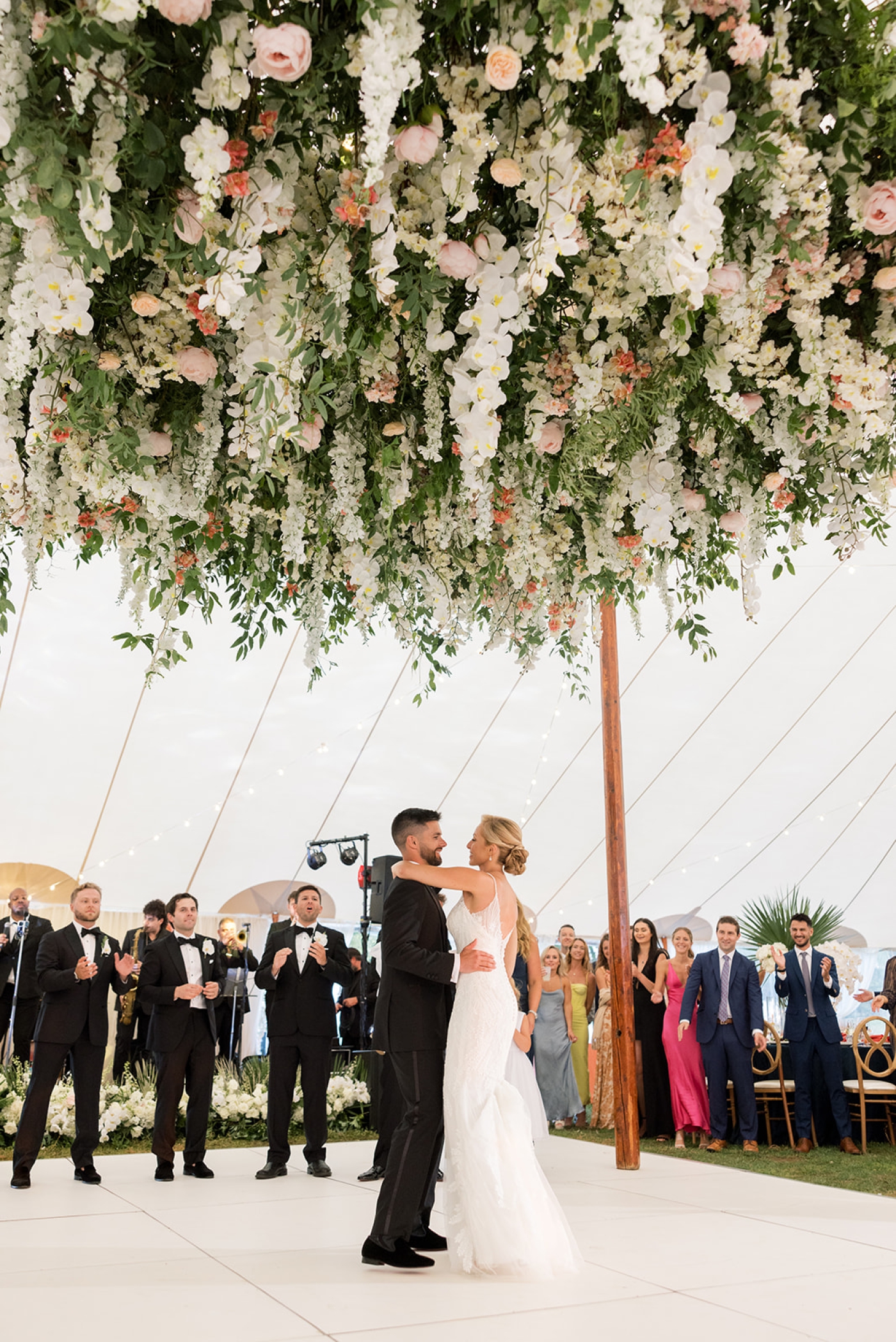 A bride and groom dance beneath a large floral installation with hanging flowers and greenery