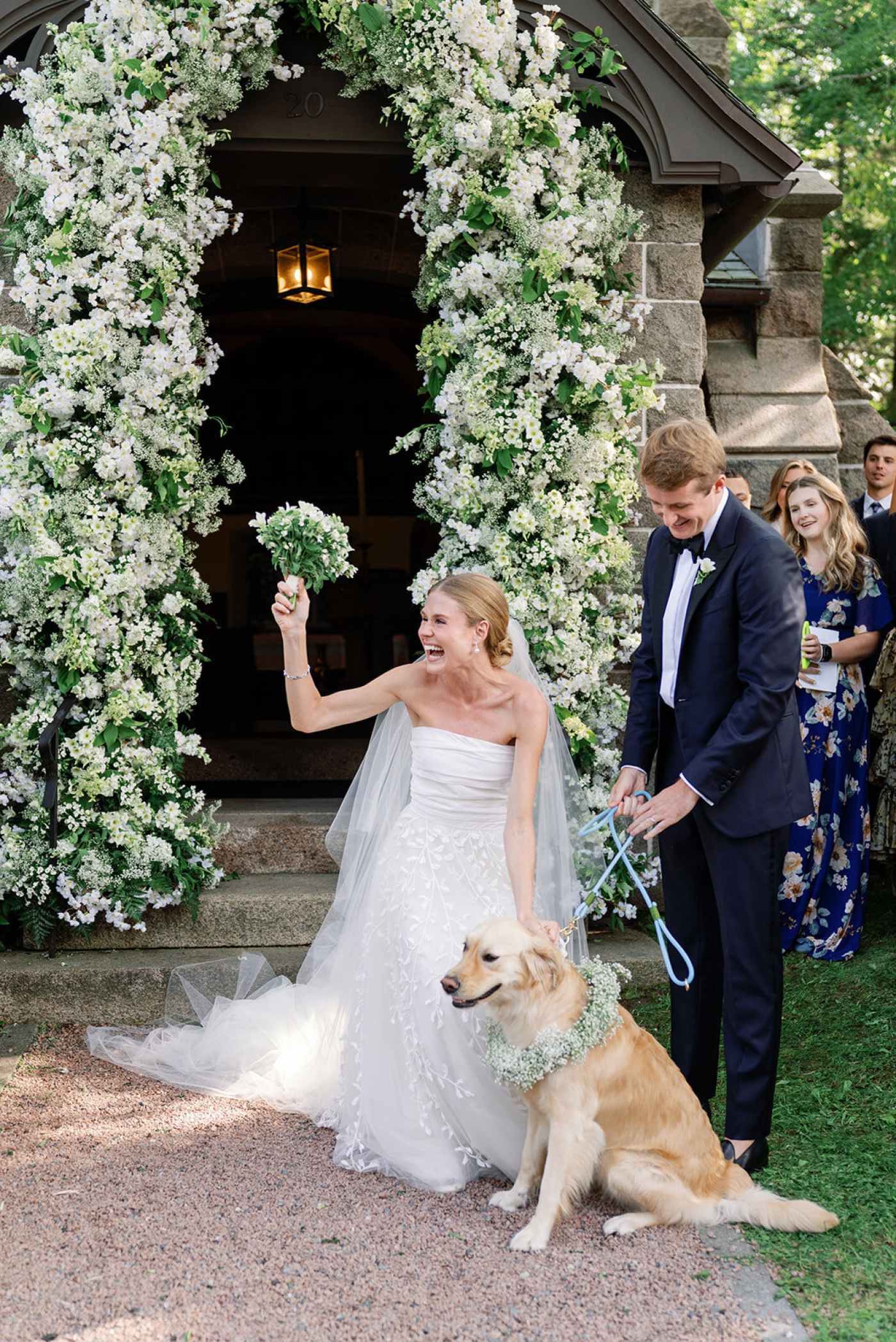 A bride and groom pet a dog while standing in front of a large floral arch made of real a faux flowers