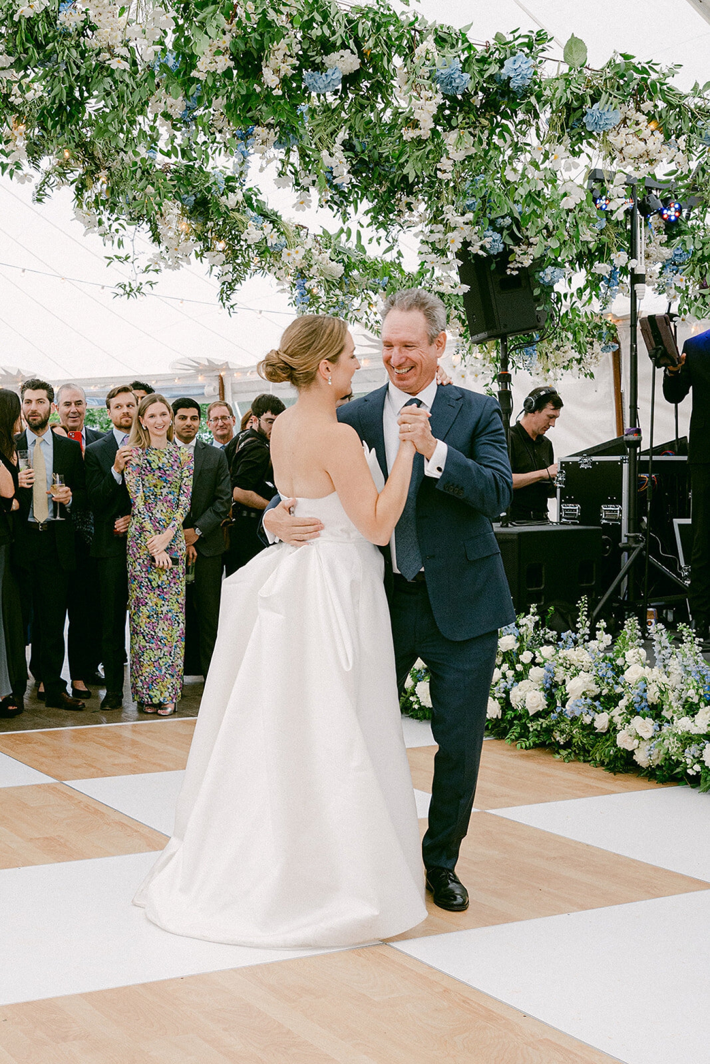 A bride dances with her dad beneath a floral chandelier made of blue, white, and green real and faux flowers