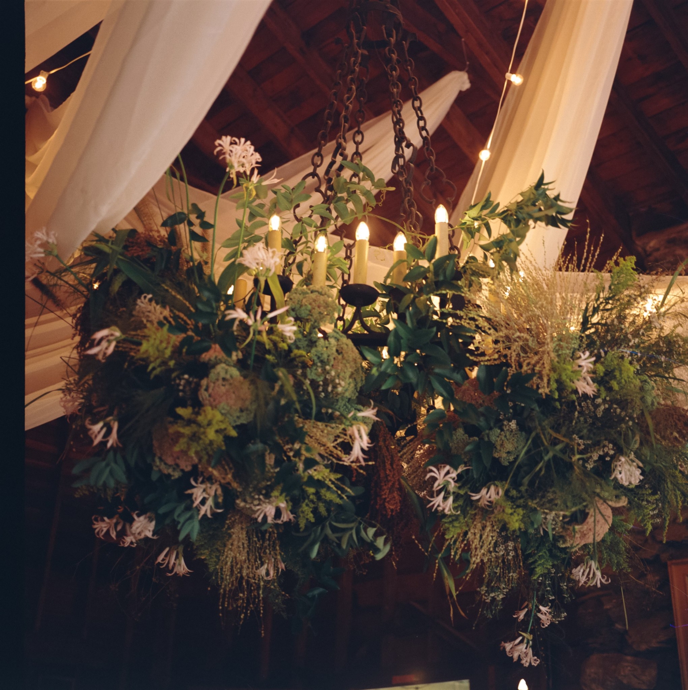 A large hanging floral installation with light blooms in dark greenery with white draping hanging from the ceiling of Megunticook Golf Club