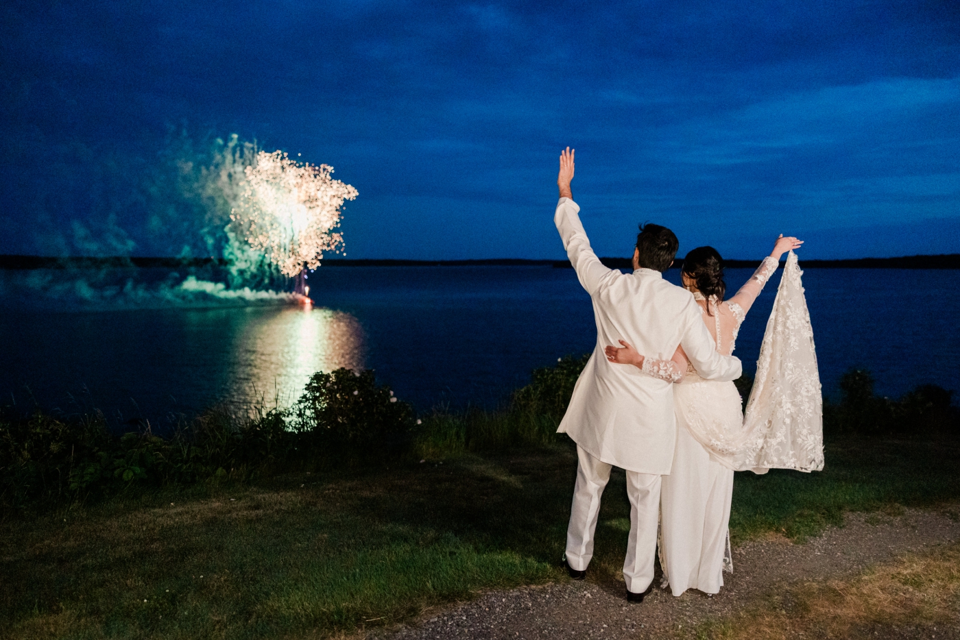 A bride and groom celebrate while fireworks go off over a lake